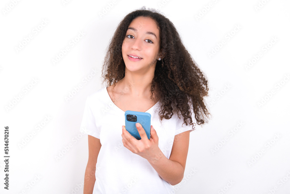 Teenager girl with afro hairstyle wearing white T-shirt over white wall holds mobile phone in hands and rejoices positive news, uses modern cellular Teenager girl with afro hairstyle wearing white T-shirt over white wall holds mobile phone in hands and rejoices positive news, uses modern cellular