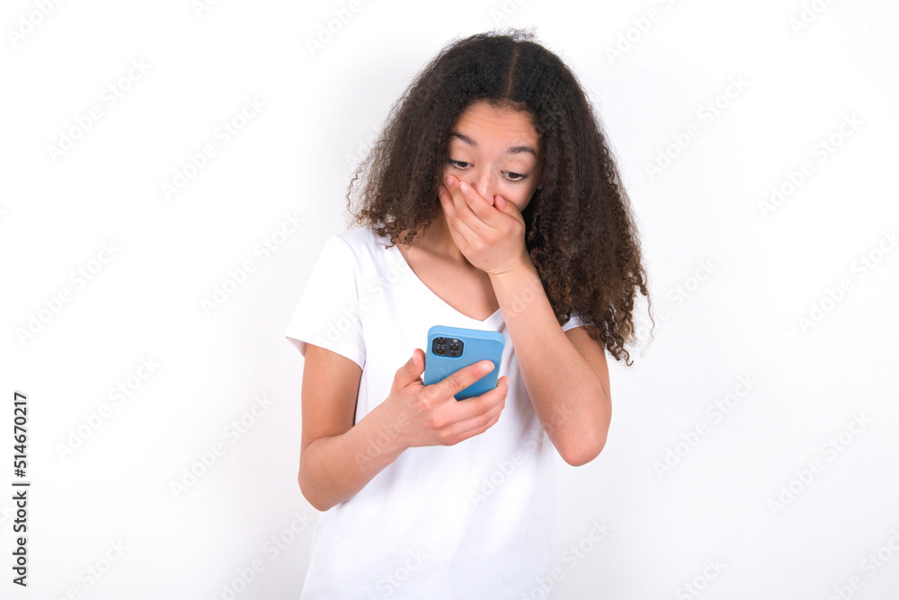 Teenager girl with afro hairstyle wearing white T-shirt over white wall being deeply surprised, stares at smartphone display, reads shocking news on website, Omg, its horrible! Teenager girl with afro hairstyle wearing white T-shirt over white wall being deeply surprised, stares at smartphone display, reads shocking news on website, Omg, its horrible!