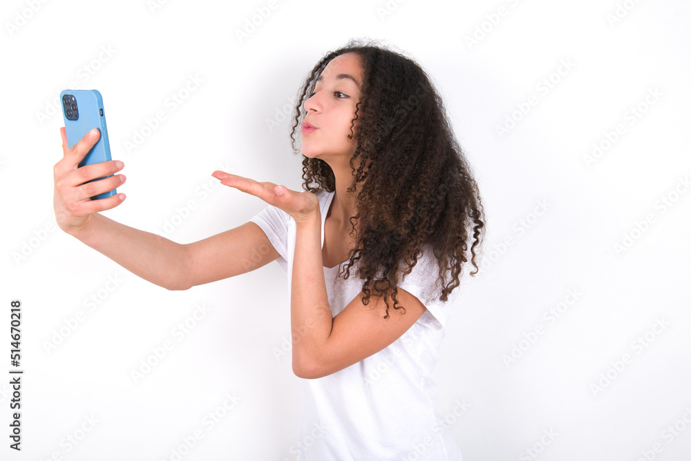 Teenager girl with afro hairstyle wearing white T-shirt over white wall blows air kiss at camera of smartphone and takes selfie, sends mwah via online call. Teenager girl with afro hairstyle wearing white T-shirt over white wall blows air kiss at camera of smartphone and takes selfie, sends mwah via online call.