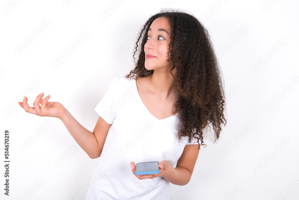 Happy pleased Teenager girl with afro hairstyle wearing white T-shirt over white wall raises palm and holds cellphone uses high speed internet for text messaging or video calls Happy pleased Teenager girl with afro hairstyle wearing white T-shirt over white wall raises palm and holds cellphone uses high speed internet for text messaging or video calls