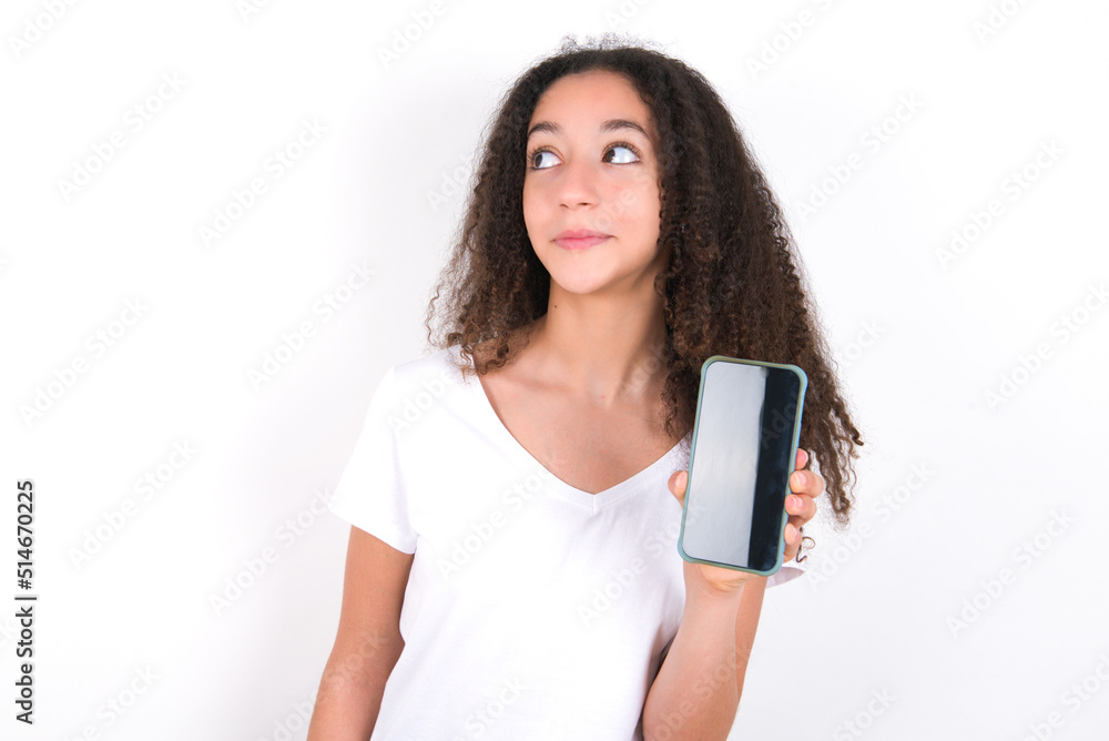 Teenager girl with afro hairstyle wearing white T-shirt over white wall holds new mobile phone and looks mysterious aside shows blank display of modern cellular Teenager girl with afro hairstyle wearing white T-shirt over white wall holds new mobile phone and looks mysterious aside shows blank display of modern cellular