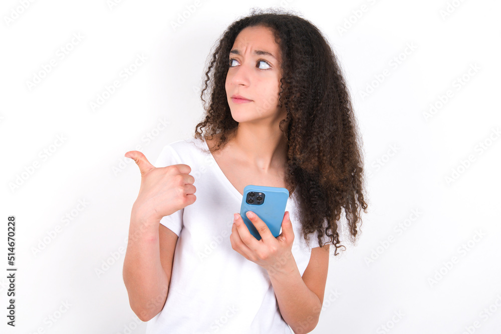 Teenager girl with afro hairstyle wearing white T-shirt over white wall points thumb away and shows blank space aside, holds mobile phone for sending text messages. Teenager girl with afro hairstyle wearing white T-shirt over white wall points thumb away and shows blank space aside, holds mobile phone for sending text messages.