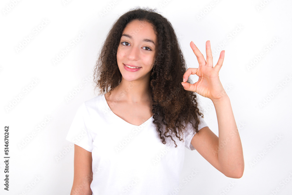 Teenager girl with afro hairstyle wearing white T-shirt over white wall hold hand arm okey symbol toothy approve advising novelty news Teenager girl with afro hairstyle wearing white T-shirt over white wall hold hand arm okey symbol toothy approve advising novelty news