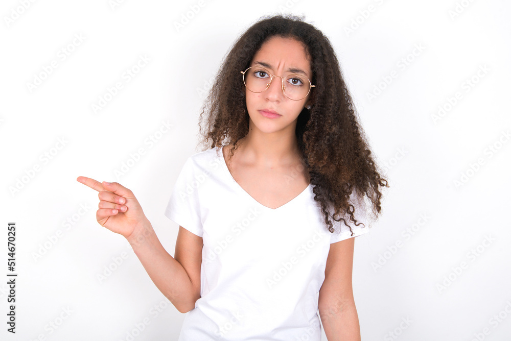 Serious Teenager girl with afro hairstyle wearing white T-shirt over white wall smirks face points away on copy space shows something unpleasant. Look at this advertisement. Big price concept Serious Teenager girl with afro hairstyle wearing white T-shirt over white wall smirks face points away on copy space shows something unpleasant. Look at this advertisement. Big price concept