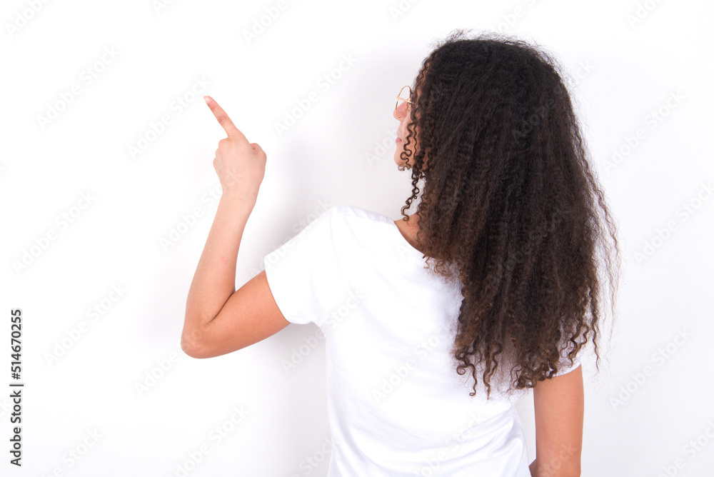 Teenager girl with afro hairstyle wearing white T-shirt over white wall pointing to object on copy space, rear view. Turn your back Teenager girl with afro hairstyle wearing white T-shirt over white wall pointing to object on copy space, rear view. Turn your back