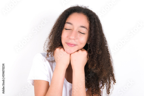 Cheerful Teenager girl with afro hairstyle wearing white T-shirt over white wall has shy satisfied expression, smiles broadly, shows white teeth, People emotions