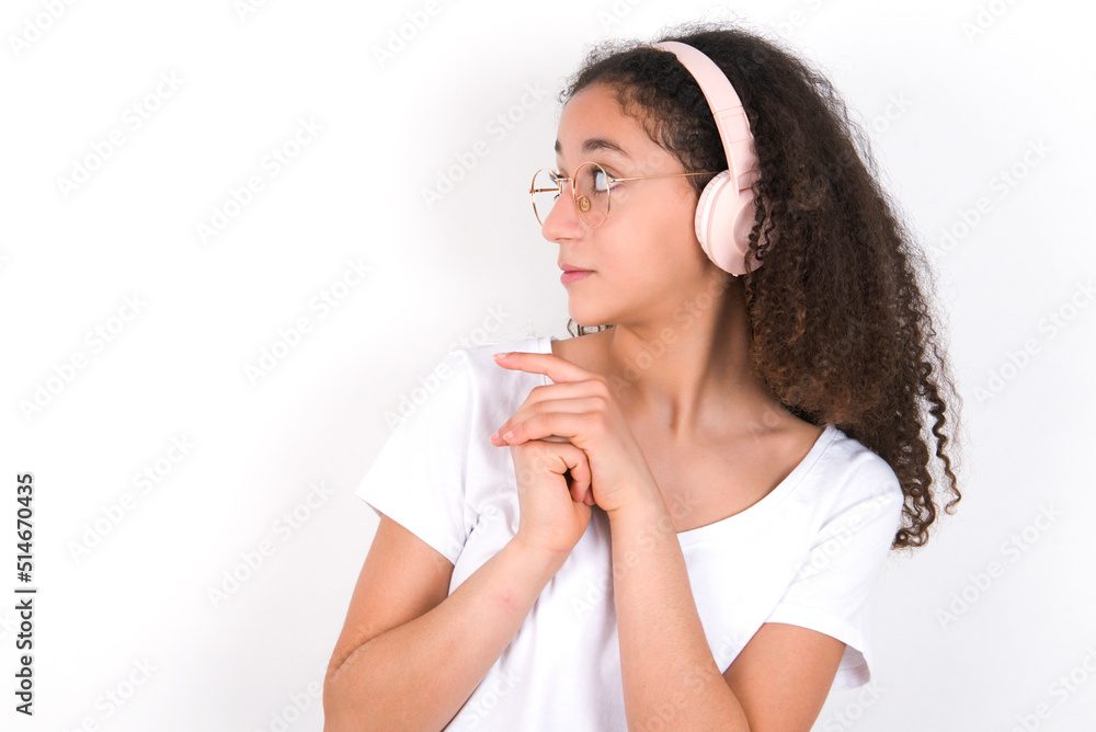 Teenager girl with afro hairstyle wearing white T-shirt over white wall wears stereo headphones listening to music concentrated and looking aside with interest. Teenager girl with afro hairstyle wearing white T-shirt over white wall wears stereo headphones listening to music concentrated and looking aside with interest.