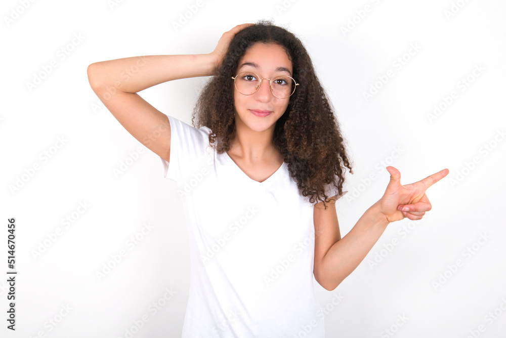 Surprised Teenager girl with afro hairstyle wearing white T-shirt over white wall pointing at empty space holding hand on head Surprised Teenager girl with afro hairstyle wearing white T-shirt over white wall pointing at empty space holding hand on head