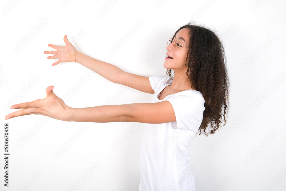 Funny astonished Teenager girl with afro hairstyle wearing white T-shirt over white wall look empty space with arms opened ready to catch something. Funny astonished Teenager girl with afro hairstyle wearing white T-shirt over white wall look empty space with arms opened ready to catch something.