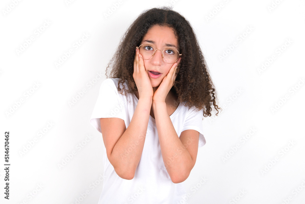 Upset Teenager girl with afro hairstyle wearing white T-shirt over white wall  touching face with two hands