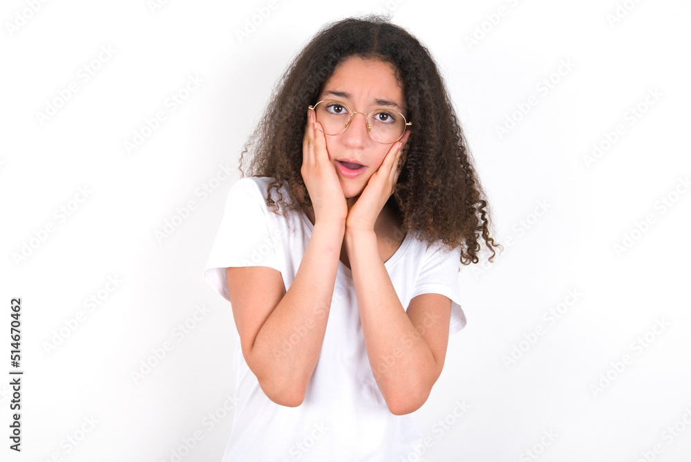 Upset Teenager girl with afro hairstyle wearing white T-shirt over white wall touching face with two hands Upset Teenager girl with afro hairstyle wearing white T-shirt over white wall touching face with two hands