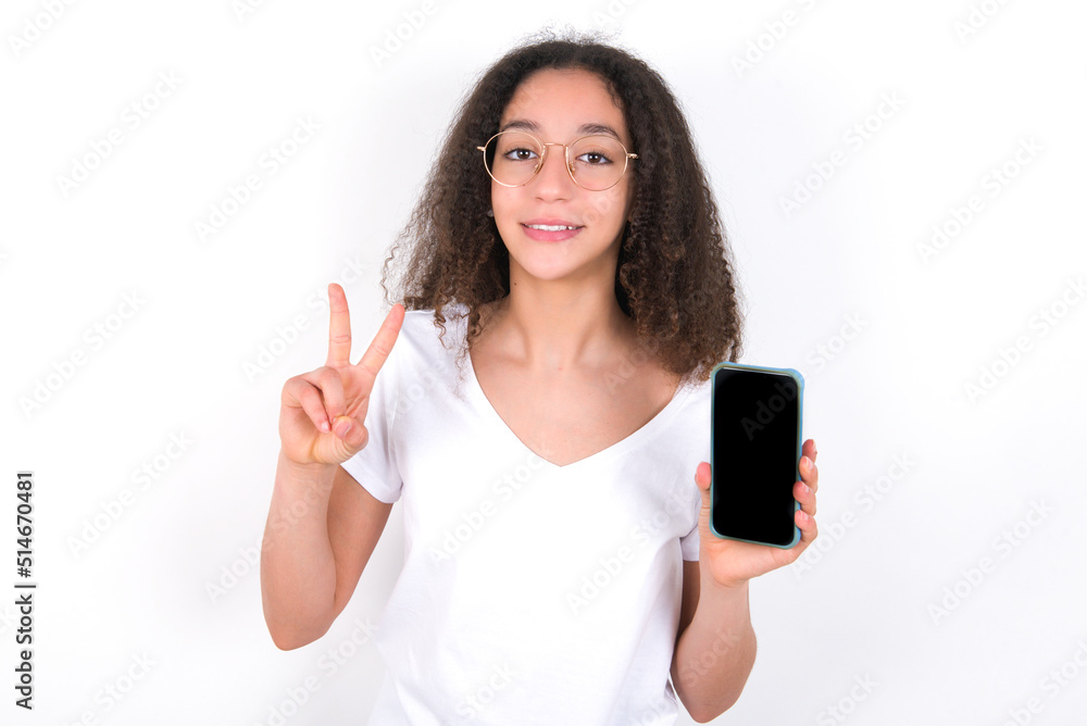Teenager girl with afro hairstyle wearing white T-shirt over white wall holding modern device showing v-sign Teenager girl with afro hairstyle wearing white T-shirt over white wall holding modern device showing v-sign