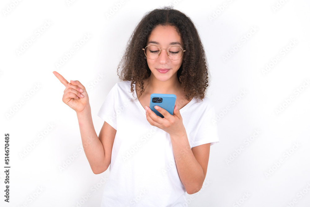 Smiling Teenager girl with afro hairstyle wearing white T-shirt over white wall pointing finger at blank space holding phone in one hand Smiling Teenager girl with afro hairstyle wearing white T-shirt over white wall pointing finger at blank space holding phone in one hand