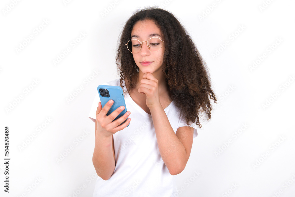 Thoughtful happy Teenager girl with afro hairstyle wearing white T-shirt over white wall hold look phone, copy space Thoughtful happy Teenager girl with afro hairstyle wearing white T-shirt over white wall hold look phone, copy space
