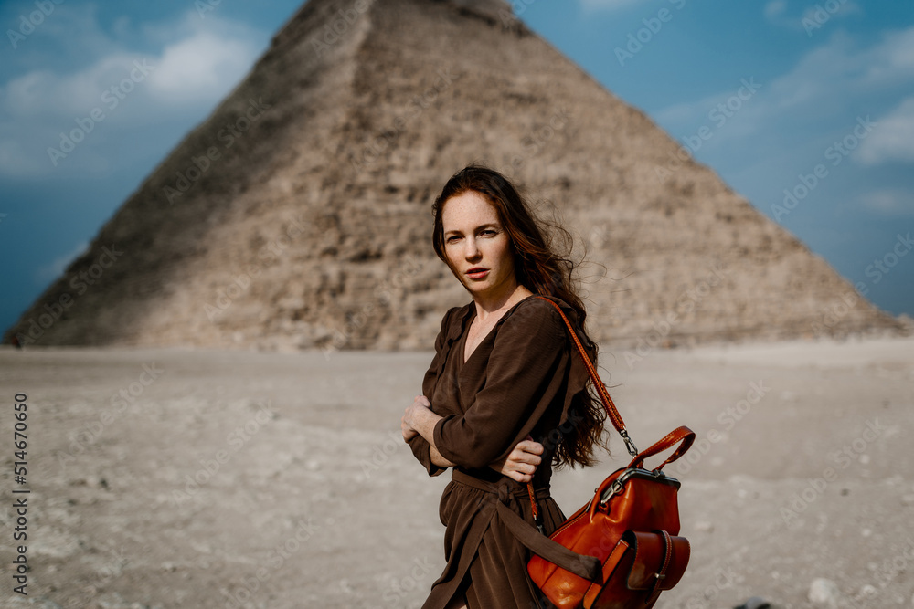 Naklejka premium Young redhead tourist girl in brawn dress with a leather backpack standing on the sand in Egypt, Cairo - Giza. Pyramids on backround. Copy space