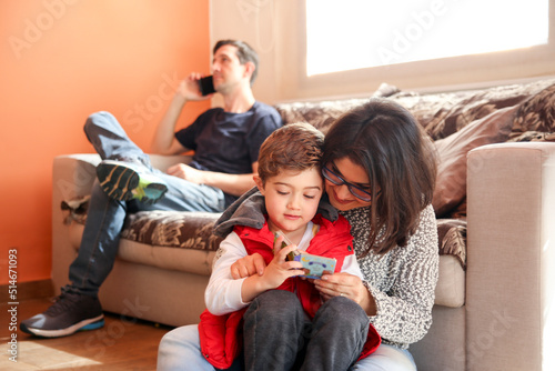 Mother Reading to Son in the Front Part of the Image, While Father is Unfocused Using a Smartphone in the Background.