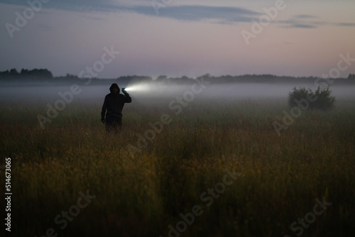 An unrecognizable man in the fog in the evening on a meadow shines with a flashlight. Mystical and mysterious atmosphere