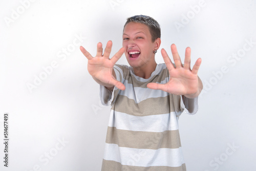 Dissatisfied young woman with short hair wearing striped t-shirt over white background frowns face, has disgusting expression, shows tongue, expresses non compliance, irritated with somebody. photo