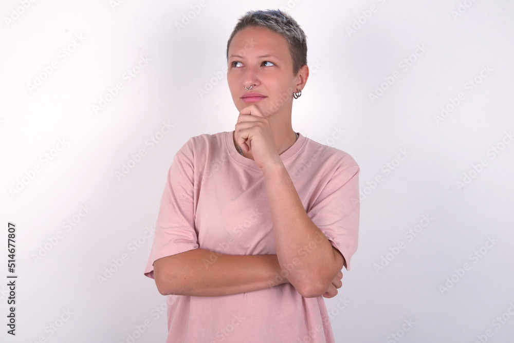 Thoughtful young woman with short hair wearing pink t-shirt over white background holds chin and looks away pensively makes up great plan Thoughtful young woman with short hair wearing pink t-shirt over white background holds chin and looks away pensively makes up great plan