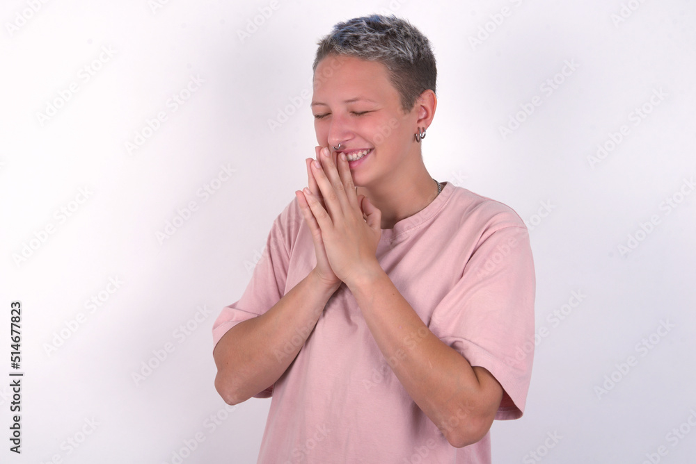 Overjoyed young woman with short hair wearing pink t-shirt over white background laughs joyfully and keeps palms pressed together hears something funny Overjoyed young woman with short hair wearing pink t-shirt over white background laughs joyfully and keeps palms pressed together hears something funny