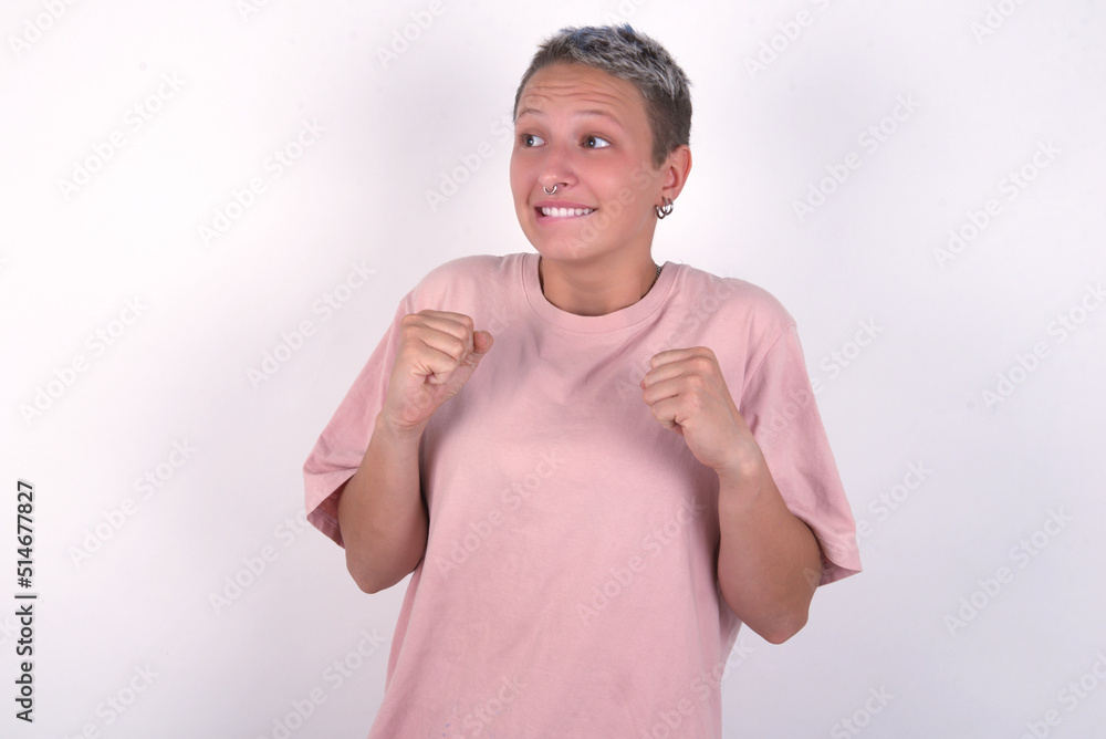 young woman with short hair wearing pink t-shirt over white background clenches fists and awaits for something nice happened looks away bites lips and waits announcement of results young woman with short hair wearing pink t-shirt over white background clenches fists and awaits for something nice happened looks away bites lips and waits announcement of results