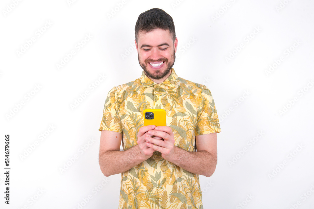 Smiling young handsome bearded man wearing flowered shirt over white wall using cell phone, messaging, being happy to text with friends, looking at smartphone. Modern technologies and communication. Smiling young handsome bearded man wearing flowered shirt over white wall using cell phone, messaging, being happy to text with friends, looking at smartphone. Modern technologies and communication.