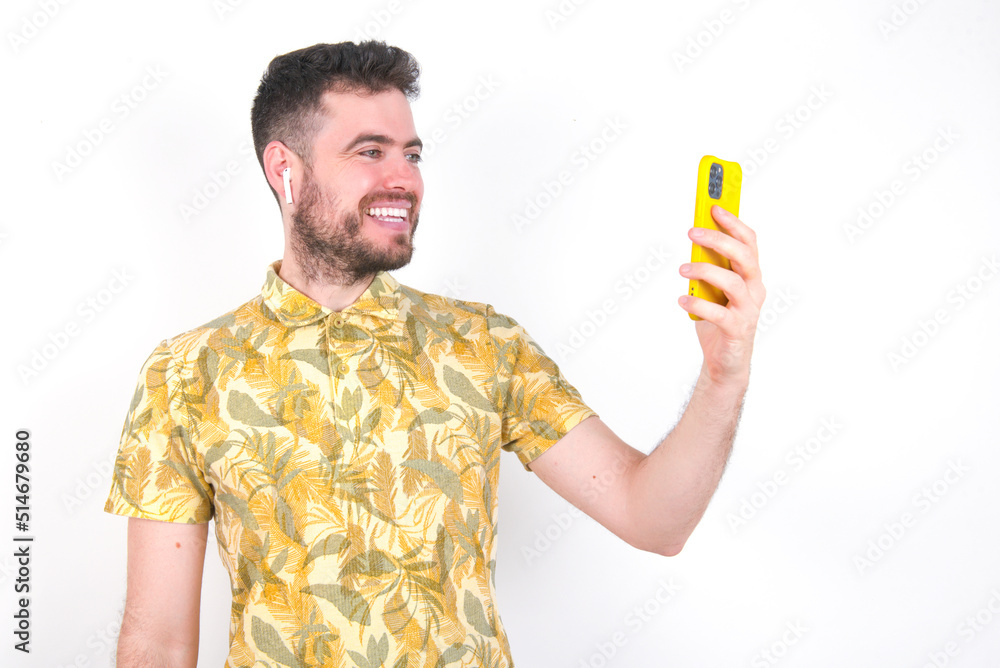 Isolated shot of pleased cheerful young handsome bearded man wearing flowered shirt over white wall, makes selfie with mobile phone. People, technology and leisure concept Isolated shot of pleased cheerful young handsome bearded man wearing flowered shirt over white wall, makes selfie with mobile phone. People, technology and leisure concept