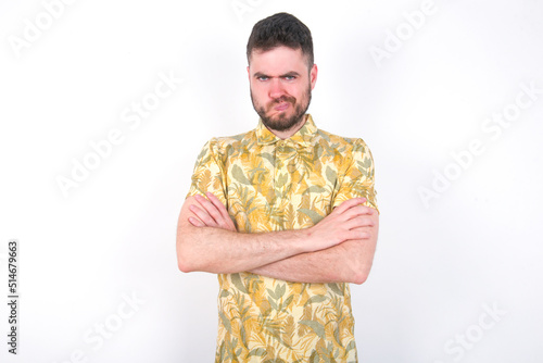 Picture of angry young handsome bearded man wearing flowered shirt over white wall crossing arms. Looking at camera with disappointed expression.
