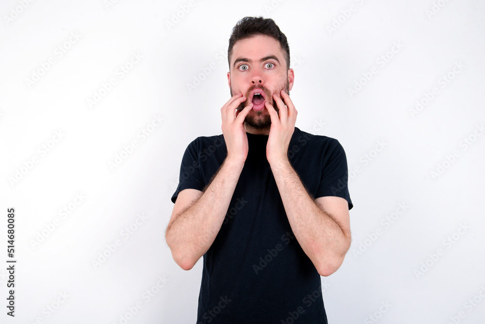 Speechless young caucasian bearded man wearing black t-shirt standing over white wall keeps hands near opened mouth reacts to shocking news stares wondered at camera Speechless young caucasian bearded man wearing black t-shirt standing over white wall keeps hands near opened mouth reacts to shocking news stares wondered at camera