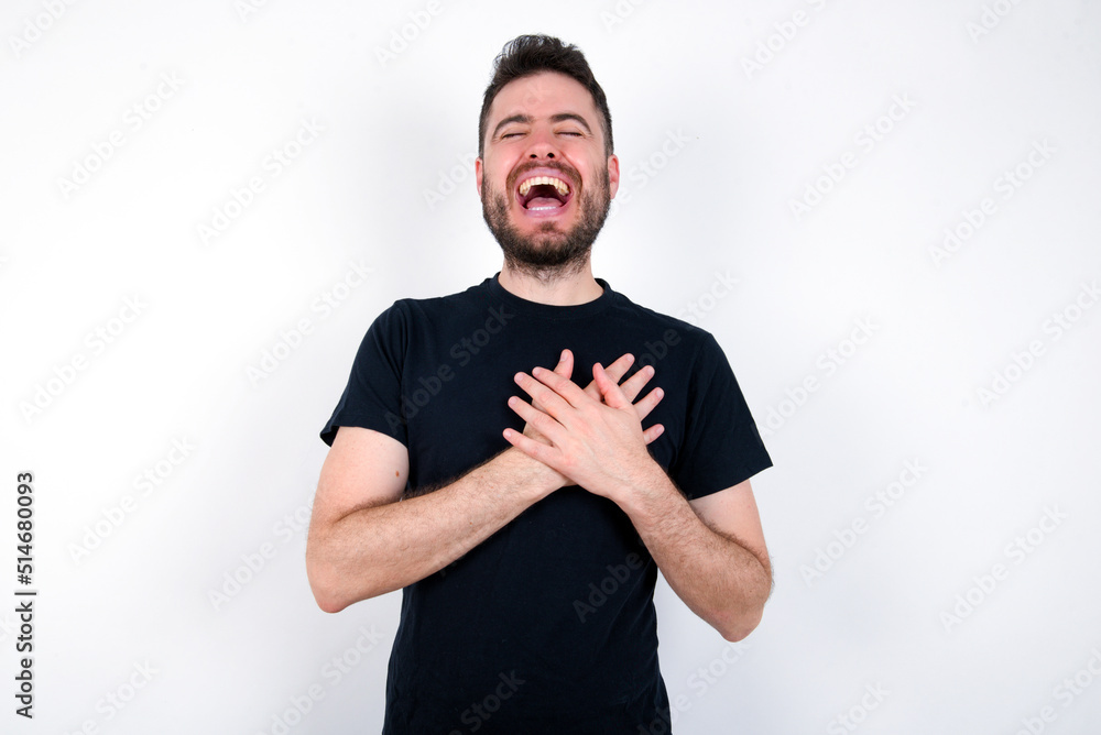 young caucasian bearded man wearing black t-shirt standing over white wall expresses happiness, laughs pleasantly, keeps hands on heart young caucasian bearded man wearing black t-shirt standing over white wall expresses happiness, laughs pleasantly, keeps hands on heart