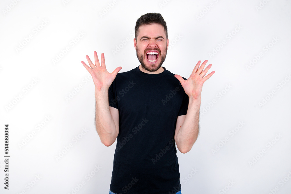 Fototapeta premium Crazy outraged young caucasian bearded man wearing black t-shirt standing over white wall screams loudly and gestures angrily yells furiously. Negative human emotions feelings concept