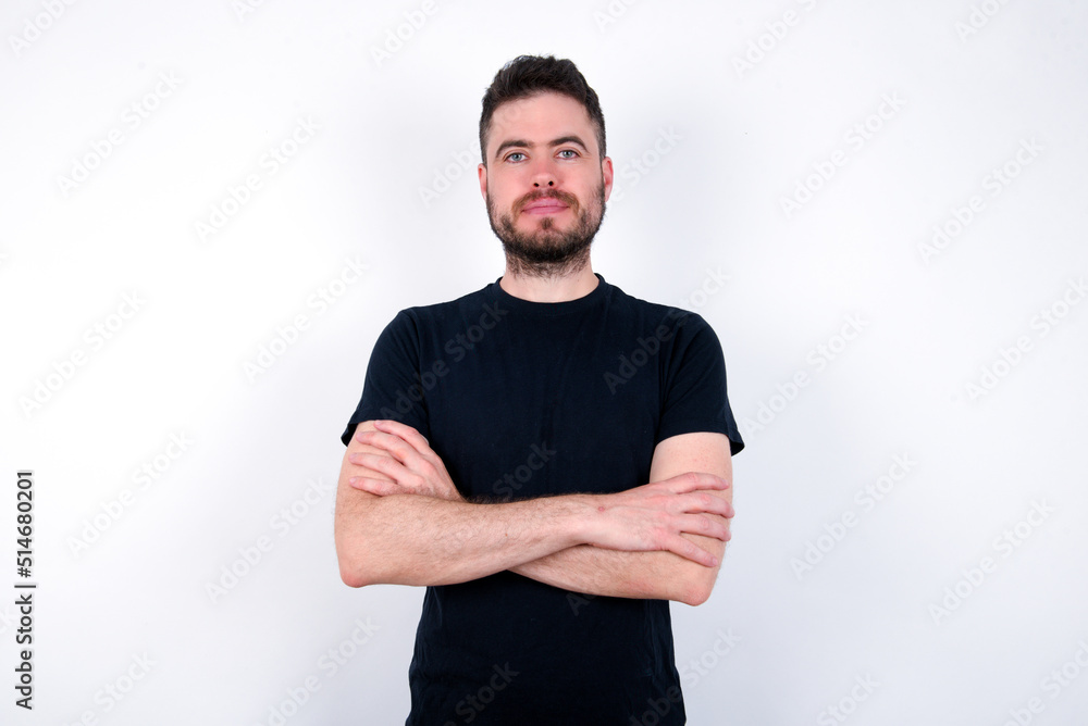 Self confident serious calm young caucasian bearded man wearing black t-shirt standing over white wall stands with arms folded. Shows professional vibe stands in assertive pose. Self confident serious calm young caucasian bearded man wearing black t-shirt standing over white wall stands with arms folded. Shows professional vibe stands in assertive pose.
