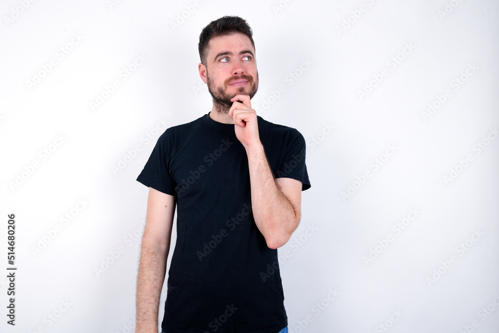 Thoughtful young caucasian bearded man wearing black t-shirt standing over white wall holds chin and looks away pensively makes up great plan Thoughtful young caucasian bearded man wearing black t-shirt standing over white wall holds chin and looks away pensively makes up great plan