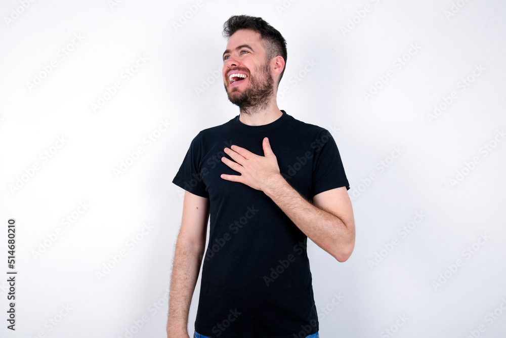Joyful young caucasian bearded man wearing black t-shirt standing over white wall expresses positive emotions recalls something funny keeps hand on chest and giggles happily. Joyful young caucasian bearded man wearing black t-shirt standing over white wall expresses positive emotions recalls something funny keeps hand on chest and giggles happily.