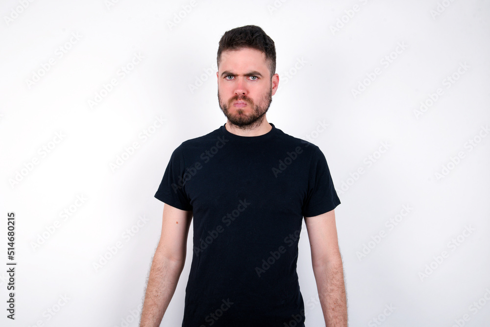 Offended dissatisfied young caucasian bearded man wearing black t-shirt standing over white wall with moody displeased expression at camera being disappointed by something Offended dissatisfied young caucasian bearded man wearing black t-shirt standing over white wall with moody displeased expression at camera being disappointed by something