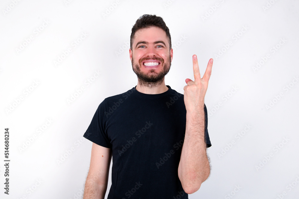 young caucasian bearded man wearing black t-shirt standing over white wall smiling and looking friendly, showing number two or second with hand forward, counting down young caucasian bearded man wearing black t-shirt standing over white wall smiling and looking friendly, showing number two or second with hand forward, counting down