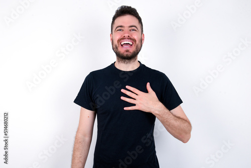 young caucasian bearded man wearing black t-shirt standing over white wall smiles toothily cannot believe eyes expresses good emotions and surprisement