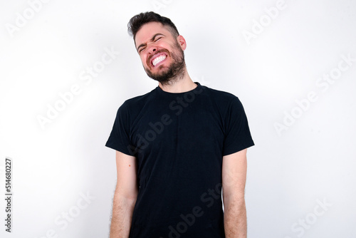 Positive young caucasian bearded man wearing black t-shirt standing over white wall with overjoyed expression closes eyes and laughs shows white perfect teeth