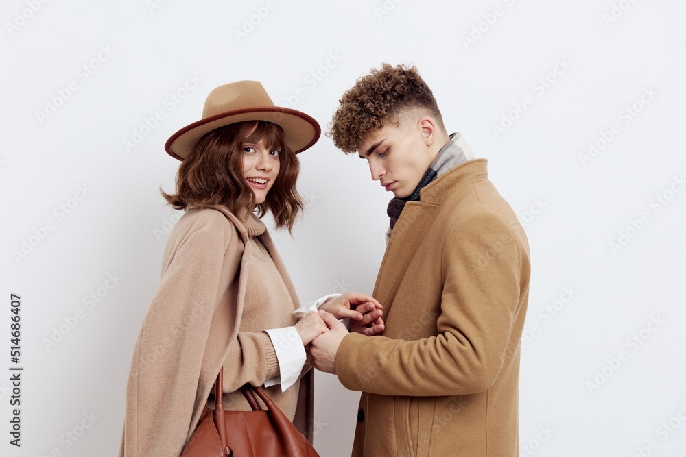 a cute couple stands on a light background dressed in coats and sweaters, a man stands against a woman and hugs her in front a cute couple stands on a light background dressed in coats and sweaters, a man stands against a woman and hugs her in front