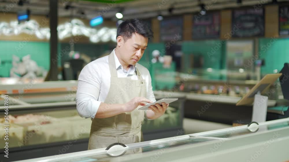 Asian grocery store manager salesman in apron using digital tablet counting goods in supermarket. Male worker employee doing inspection of products regular inventory to control goods, online order Asian grocery store manager salesman in apron using digital tablet counting goods in supermarket. Male worker employee doing inspection of products regular inventory to control goods, online order