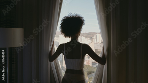 Beautiful Latin woman waking up in the morning, the sun shines on her from the big window. Happy young woman greets the new day with warm sunlight and city scenery in the window.