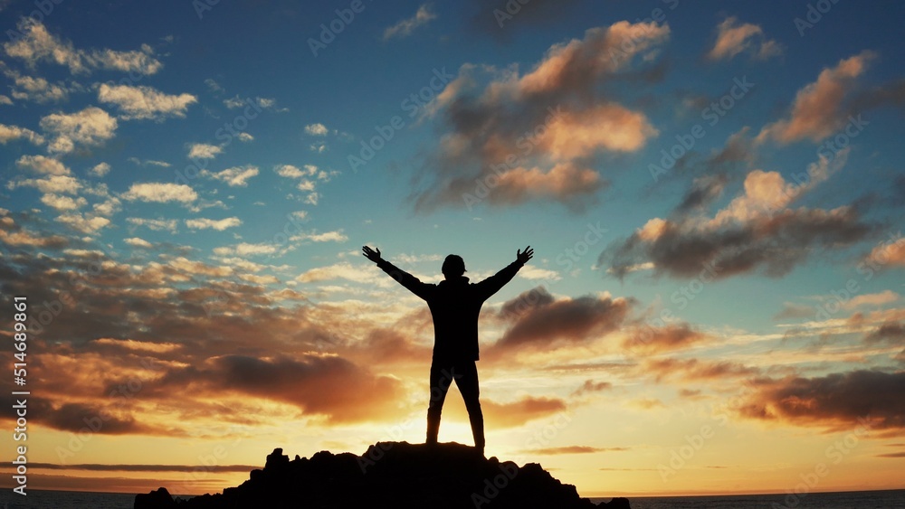 Man stands on a rock against the ocean during summer sunset. Human with raised hands looks to sun over horizon in morning while sunrise. Man feels happy and freedom. Silhouette of a man against  sky