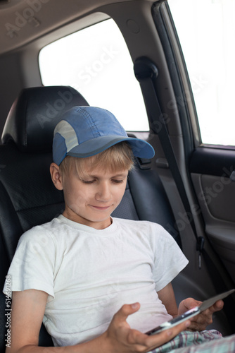 A boy in a cap sits in the back seat of a car, looks at the phone screen and smiles
