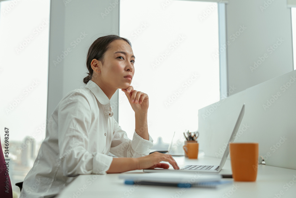 Asian business woman working laptop sitting in modern office and looking away. High quality photo Asian business woman working laptop sitting in modern office and looking away. High quality photo