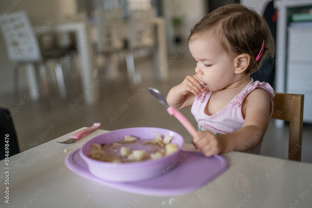 One child small caucasian toddler female baby hold spoon eating at the table alone at home in room real people copy space early child development concept learning front view hold food healthy eating One child small caucasian toddler female baby hold spoon eating at the table alone at home in room real people copy space early child development concept learning front view hold food healthy eating