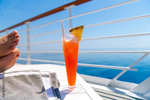 A woman rests her feet on a table on the upper deck of a cruise ship at sea with a colorful drink with pineapple on a hot summer day on the ocean.