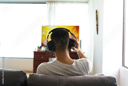 person from the back sitting on a sofa in the living room of a house, using wireless headphones to listen to music in front of television, technology and lifestyle with hobbies photo