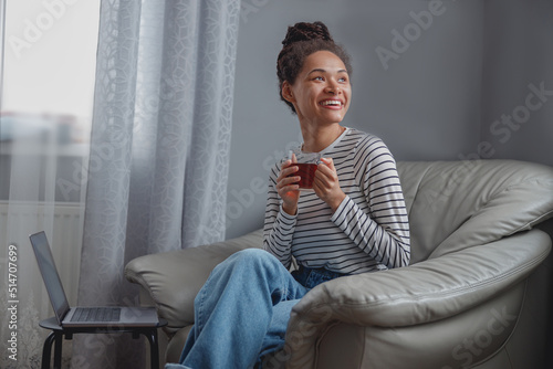 Cheerful young pretty female with braids drinking tea at laptop in room. Rest from freelance.
