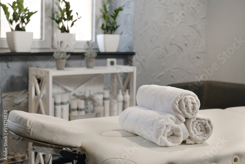 Rolled white towels on massage table in empty salon. Interior of aesthetic clinic, nobody. Modern cabinet of physiotherapist. Empty room with plants of beautician in wellness center. Copy space photo