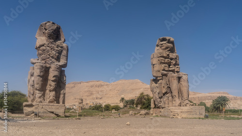 Statues of the colossi of Memnon against a clear blue sky and a sand dune. Giant sculptures of seated pharaohs are damaged. Green vegetation around. Egypt